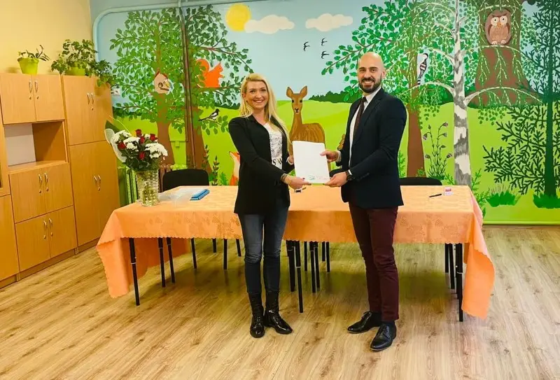 Zdjęcie o orientacji poziomej przedstawia kobietę i mężczyznę stojących w sali przedszkolnej, trzymających wspólnie biały dokument. Kobieta ma na sobie czarną marynarkę, jeansy i ciemne botki, natomiast mężczyzna jest ubrany w elegancki granatowy garnitur i bordowe spodnie. Za nimi znajduje się stół przykryty brzoskwiniowym obrusem, na którym stoi wazon z bukietem białych i czerwonych kwiatów. Ścianę zdobi kolorowy, bajkowy mural przedstawiający las, zwierzęta oraz drzewa, co nadaje wnętrzu edukacyjny i przyjazny charakter. Po lewej stronie widoczne są wysokie, drewniane szafki, a na podłodze ułożone są panele w odcieniu jasnego drewna. Całość kompozycji jest symetryczna i dobrze oświetlona, podkreślając oficjalny, a zarazem ciepły klimat spotkania.
