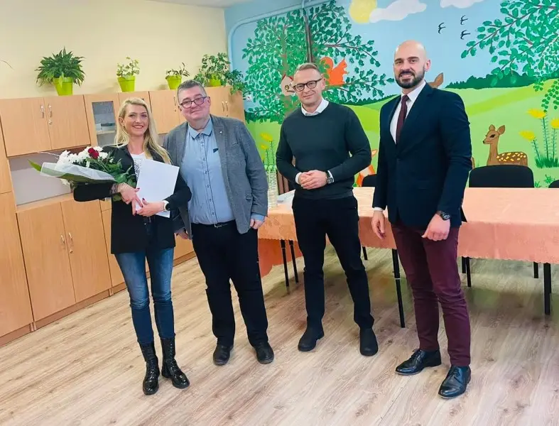 Zdjęcie o orientacji pionowej przedstawia grupę czterech osób stojących w jasnej sali przedszkolnej, prawdopodobnie podczas oficjalnego spotkania lub uroczystości. Od lewej strony widoczna jest kobieta z bukietem kwiatów i dokumentami, obok której stoją trzej mężczyźni w eleganckich strojach. W tle widać drewniane szafki oraz ścianę ozdobioną kolorowym malowidłem ściennym z motywami przyrody, takimi jak drzewa, wiewiórka i jelonek. Całość kompozycji dopełnia stół przykryty pomarańczowym obrusem oraz jasna podłoga w odcieniach drewna. Zdjęcie ma charakter formalny i pozytywny, podkreślający współpracę w otoczeniu sprzyjającym edukacji dzieci.