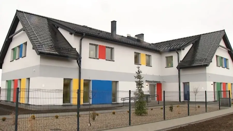Zdjęcie przedstawia nowoczesny budynek użyteczności publicznej lub przedszkole w układzie poziomym o proporcjach około 16:9. Biała elewacja budynku została urozmaicona charakterystycznymi, wielobarwnymi pionowymi panelami w kolorach czerwonym, niebieskim, zielonym i żółtym, które znajdują się pomiędzy oknami. Całość zwieńczona jest ciemnym, dwuspadowym dachem z dachówki, a na środkowej części budynku widoczny jest szary pas dzielący kondygnacje. Teren wokół obiektu jest ogrodzony ażurowym, metalowym płotem, za którym widać żwirowe podłoże oraz młode nasadzenia drzewek. Wejście do budynku z podjazdem dla osób niepełnosprawnych znajduje się w centralnej części, a oświetlenie zewnętrzne podkreśla nowoczesny charakter placówki.